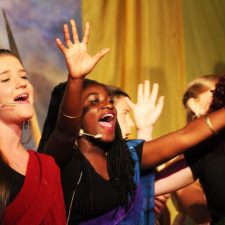 Chorus of women singing with cheek microphones, in costume